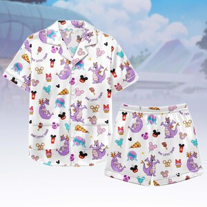 May include: White pyjama set featuring a repeating pattern of a purple dragon, Mickey Mouse, pizza slices, and other icons. The short-sleeved top and shorts have white trim. The text "ONE LITTLE SPARK" is also visible.