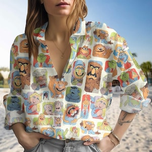 May include: A long-sleeved shirt with a repeating pattern of Muppet characters in various poses and colours. The shirt has a V-neck and is made of a light-coloured fabric.