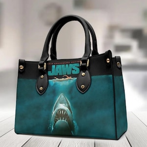 Op de afbeelding: Zwarte en blauwgroene handtas met de filmposter van "Jaws". De tas heeft zwarte handvatten en afwerking, met goudkleurige hardware. De poster toont een haai onder water met de filmtitel bovenaan.