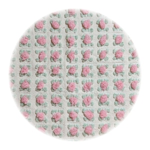 May include: A round, white rug with a repeating pattern of pink floral tufts and light green square accents. The rug has a soft, textured appearance, suitable for a bedroom or living space.