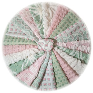 May include: A circular textile piece composed of radiating triangular fabric segments in shades of sage green, soft pink, and cream white. The fabric features varied textures including raised dots, cable knit patterns, and floral motifs.