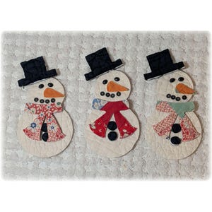 May include: Three handmade snowman ornaments. Each snowman has a black top hat, a carrot nose, and button eyes. They wear patterned scarves in red, blue, and green. The snowmen are on a textured, off-white background.