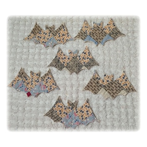 May include: A collection of five fabric bat-shaped ornaments arranged in a cluster. Each bat is constructed from various patterned textiles featuring geometric, floral, and striped designs in shades of blue, tan, cream, and grey with stitched edges.