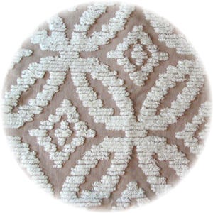 May include: A round rug with a light brown background and a white, textured geometric pattern. The design features repeating diamond and zigzag shapes, creating a soft, inviting texture. The rug is suitable for home decor.