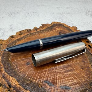 Puede incluir: Una pluma estilográfica azul marino con capuchón y adornos de metal plateado. La pluma está sobre una superficie de madera con un patrón de vetas natural. El capuchón está separado, mostrando el plumín. Aproximadamente 14 cm de largo.