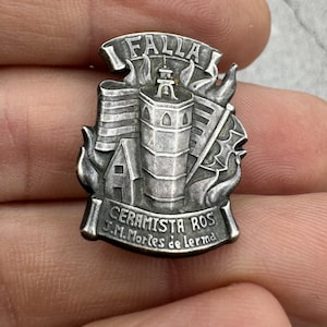 May include: A silver-toned metal pin featuring a detailed design of a building with flames and the text "FALLA CERAMISTA ROS J.M.Morles de Lerma".