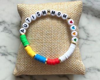 May include: A colorful beaded bracelet with the words "AUTISM MOM" spelled out in white letter beads. The bracelet features a rainbow of colors, including green, yellow, blue, red, and white, with gold accents and heart-shaped beads.