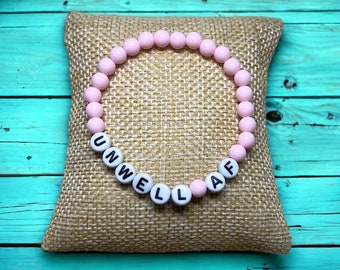 Puede incluir: Un brazalete de cuentas rosadas con la palabra "UNWELL AF" escrita en letras negras.