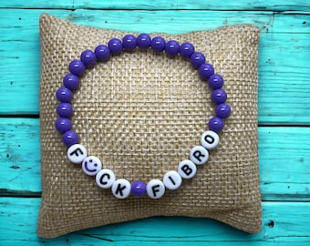Puede incluir: Pulsera de cuentas moradas con cuentas blancas que deletrean "F**K FIBRO" con una carita sonriente en la "K". La pulsera está sobre una almohada de arpillera marrón.