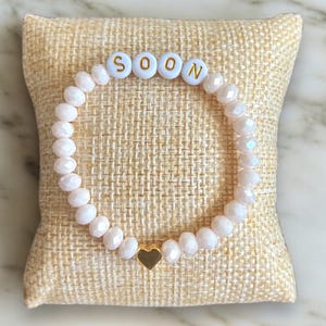 May include: A beaded bracelet with the word "SOON" spelled out in white letter beads. The bracelet features light pink faceted beads and a gold heart charm. The bracelet is displayed on a beige, textured pillow.