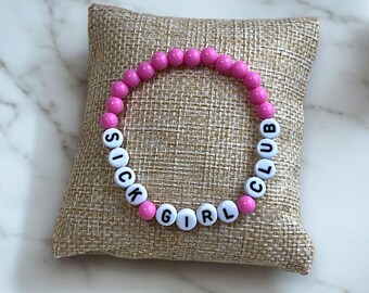 Puede incluir: Pulsera de cuentas rosadas con cuentas de letras blancas que deletrean "Sick Girl Club".
