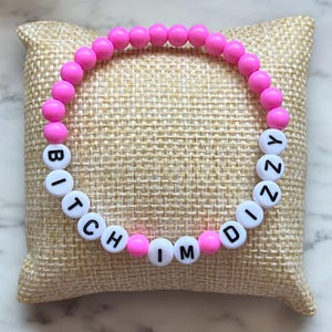 Puede incluir: Una pulsera de cuentas rosas con cuentas de letras blancas que deletrean "BITCH MINDY". La pulsera está hecha de pequeñas cuentas rosas redondas ensartadas con cuentas de letras blancas. La pulsera se muestra sobre una superficie beige texturizada.