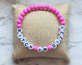 Puede incluir: Una pulsera de cuentas rosas con cuentas de letras blancas que deletrean "BITCH MINDY". La pulsera está hecha de pequeñas cuentas rosas redondas ensartadas con cuentas de letras blancas. La pulsera se muestra sobre una superficie beige texturizada.
