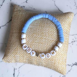 May include: A light blue beaded bracelet with the words "BOY MOM" spelled out in white letter beads. The bracelet has gold accents and is resting on a beige cushion.