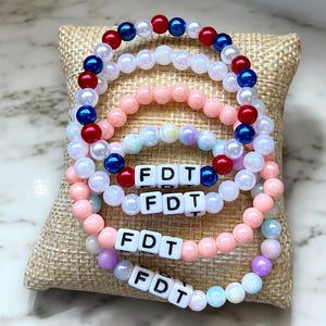 May include: Four beaded bracelets with the letters "FDT" spelled out on each. The bracelets are layered on top of each other, with the top bracelet being red, white, and blue. The other bracelets are pink, white, and light blue.