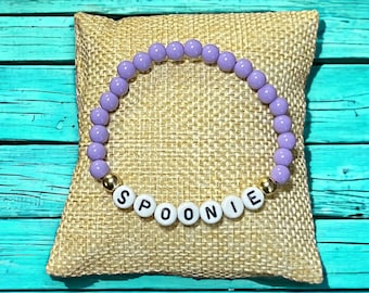 Puede incluir: Un brazalete de cuentas moradas con separadores dorados y cuentas de letras blancas que deletrean "SPOONIE".