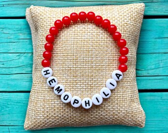 Op de afbeelding: Een rode kralenarmband met de tekst "HEMOPHILIA" met witte letterkralen. De armband ligt op een beige jute kussen tegen een turquoise houten achtergrond. De kralen zijn rond en glanzend.