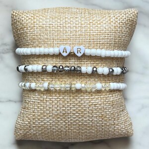 Pode incluir: Três pulseiras de contas brancas empilhadas em um travesseiro de juta bege. A pulseira superior tem duas contas brancas em forma de coração com as letras "A" e "R" em ouro.