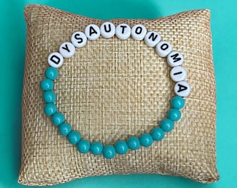 May include: A turquoise beaded bracelet with white letter beads spelling out "Dysautonomia". The bracelet is displayed on a brown burlap cushion.