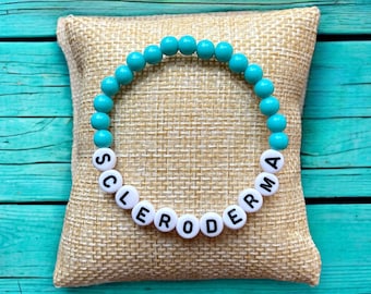 Puede incluir: Un brazalete de cuentas turquesa con cuentas de letras blancas que deletrean "SCLERODERMA".