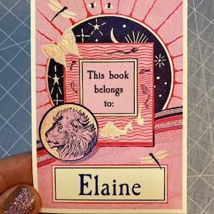 A hand holds a vintage bookplate in pink, red, navy blue and pale yellow features Art Deco-style imagery including a lion, butterflies, fish, dragonflies, and sun, moon, stars. Bookplate text is in dark blue