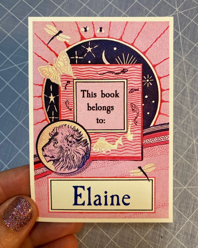 A hand holds a vintage bookplate in pink, red, navy blue and pale yellow features Art Deco-style imagery including a lion, butterflies, fish, dragonflies, and sun, moon, stars. Bookplate text is in dark blue