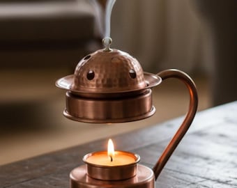 May include: A copper incense burner with a candle burning below and smoke rising from the top. The burner has a domed lid with small holes and a curved handle. The candle is in a small, round holder.