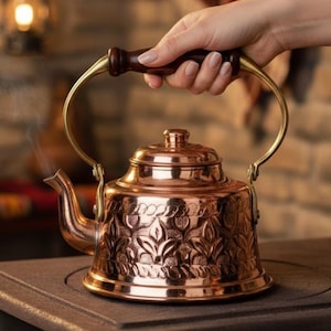 Könnte beinhalten: Eine kupferne Teekanne mit floralem Muster wird gehalten. Sie hat einen dunklen Holzgriff und einen gebogenen Messinggriff. Dampf steigt aus dem Ausguss auf. Die Teekanne steht auf einer dunklen Oberfläche.