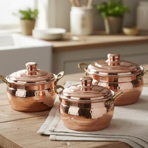 May include: Three copper cooking pots with lids and brass handles are arranged on a wooden table. The pots have a hammered texture and a shiny finish. The lids have a small knob on top. The pots are sitting on a light gray linen napkin.