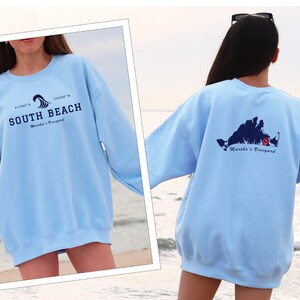 Könnte beinhalten: Hellblauer Pullover mit einem Grafikdruck einer Welle und dem Text "SOUTH BEACH Martha's Vineyard" auf der Vorderseite und einer Karte von Martha's Vineyard mit einer roten Stecknadel auf der Rückseite.