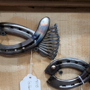 May include: Two metal fish sculptures crafted from horseshoes and other metal pieces. The fish have a dark, metallic finish with a hint of color on the fins. Each fish is mounted on a wooden surface.