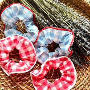 May include: Four gingham scrunchies in red and blue, with white and red trim. The scrunchies are arranged on a woven basket with dried lavender sprigs in the background. The scrunchies are a hair accessory.
