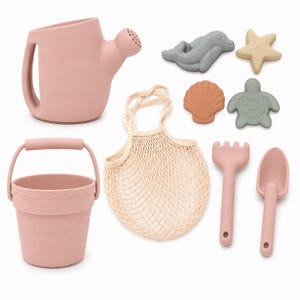 May include: A collection of beach toys in soft pastel colors. Includes a pink watering can, bucket, rake, and shovel. Also features a gray dolphin, starfish, turtle, and seashell-shaped molds, and a beige mesh bag.