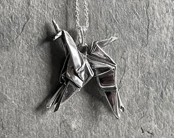 May include: A silver origami crane pendant hangs from a delicate chain. The pendant is intricately folded, showcasing the detailed craftsmanship of the paper crane design. The necklace is displayed against a textured gray background.