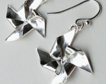 May include: A pair of silver earrings shaped like pinwheels. The earrings have a shiny, metallic finish and are suspended from simple silver hooks.