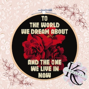 Könnte beinhalten: Kreuzstich-Stickerei in einem Holzreifen. Das Design zeigt einen schwarzen Hintergrund mit roten Rosen und dem Text "TO THE WORLD WE DREAM ABOUT AND THE ONE WE LIVE IN NOW". Der Hintergrund ist hellrosa mit floralen Mustern.