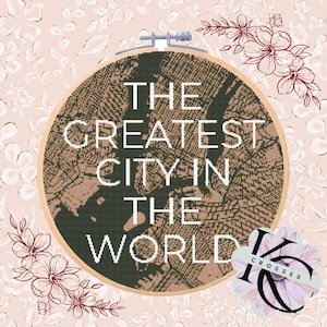 May include: Cross-stitch embroidery in a wooden hoop with the text "THE GREATEST CITY IN THE WORLD." The design features a detailed pattern in shades of green and tan. The background is a floral pattern in pink and gold.
