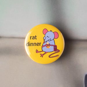 May include: A yellow button with the words "rat dinner" and a cartoon mouse holding cheese. The mouse is blue with pink ears and feet. The button has a red tail and a small red container.