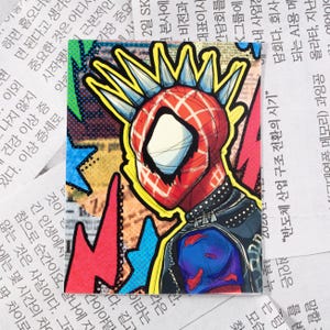 May include: A colourful sticker featuring a cartoon illustration of a superhero with a red and black suit, a mohawk, and a black leather jacket. The background is a collage of colourful comic book panels.