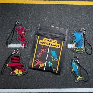 May include: A set of five colorful, cartoon-style keychains featuring a rabbit, a rat, a fish, and two variations of a cat. The keychains are made of clear plastic and have black string attachments. The keychains are packaged in a black zippered bag with the text "Roadkill Mystery Bags".