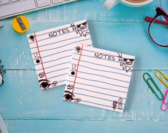 May include: Two white lined paper note pads with the word "NOTES" and a cartoon sun with sunglasses. The pads have a red vertical line and black hole punches. The pads are on a light blue wooden surface with office supplies.