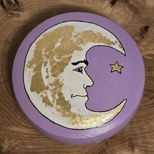 May include: A round, lavender-colored wooden disc featuring a crescent moon with a face and a single star. The moon is white with gold leaf accents, and the star is gold. The moon and star are outlined in black.