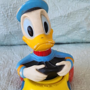 May include: A yellow toy car with a blue and red steering wheel. A cartoon duck wearing a blue hat is sitting in the driver's seat.