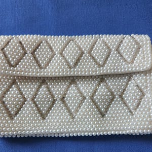 May include: A white beaded clutch purse with a diamond pattern. The purse is made of small, white pearls.
