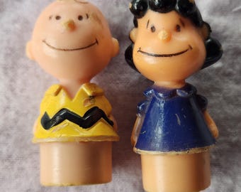 May include: Two small plastic character figurines. One figure has a bald head and wears a yellow shirt with a black zigzag pattern. The other figure has black hair and wears a dark blue dress. Both figures have peach-colored skin and painted facial features.