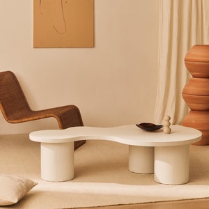 Op de afbeelding: Witte organisch gevormde salontafel met drie poten. De tafel heeft een glad, wit oppervlak en is gemaakt van een natuurlijk materiaal. De tafel staat in een woonkamer met een rieten stoel en een bruine muur.