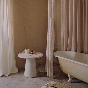 Peut inclure: Une salle de bain blanche avec une baignoire blanche, un rideau de douche blanc et une table d'appoint blanche avec un rouleau de papier toilette et une bouteille de savon liquide. Les murs sont de couleur beige clair.