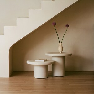 Peut inclure: Deux tables d'appoint rondes blanches avec un livre sur la plus petite table. Un vase avec deux fleurs violettes est posé sur la plus grande table. Les tables sont devant un escalier blanc.