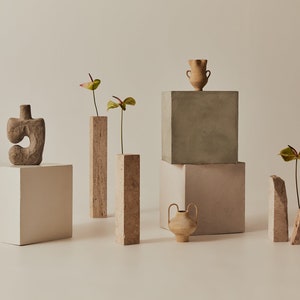 Peut inclure: Une nature morte minimaliste avec des vases et des sculptures en céramique. Les objets sont disposés sur des plateformes cubiques blanches et grises. Les vases sont remplis de fleurs vertes et roses.