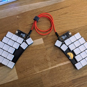Può includere: Due metà di tastiera ergonomica in bianco e nero con tasti bianchi e un cavo rosso che li collega. Le metà della tastiera sono su una superficie di legno.
